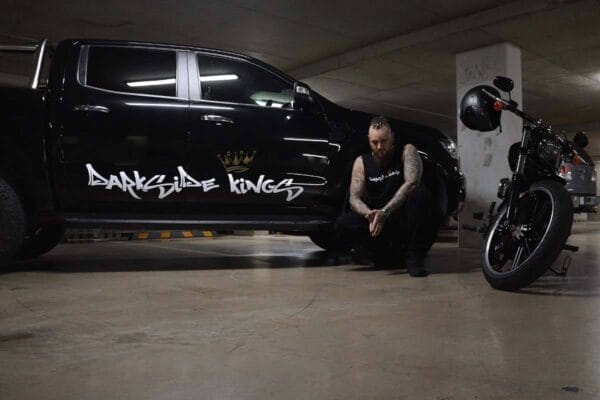 Front view of a male wearing a DSK Singlet crouching next to motorcycle and Ford Ranger