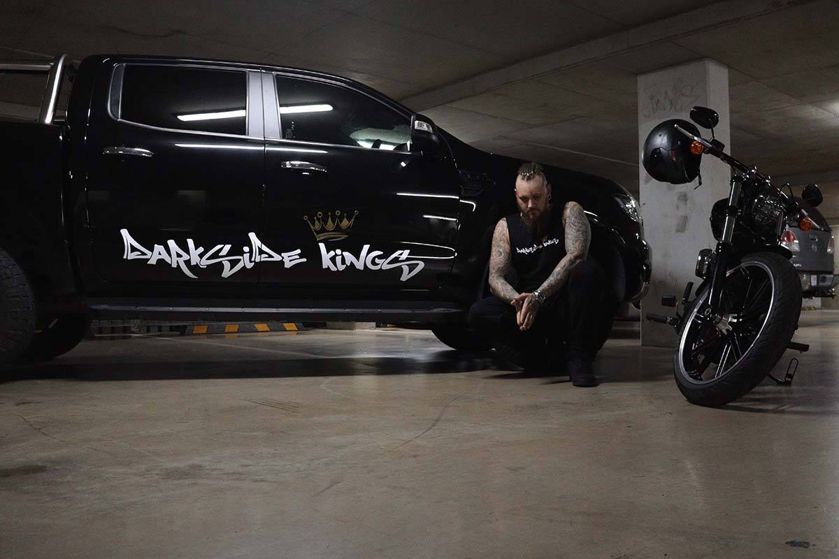 Front view of a male wearing a DSK Singlet crouching next to motorcycle and Ford Ranger