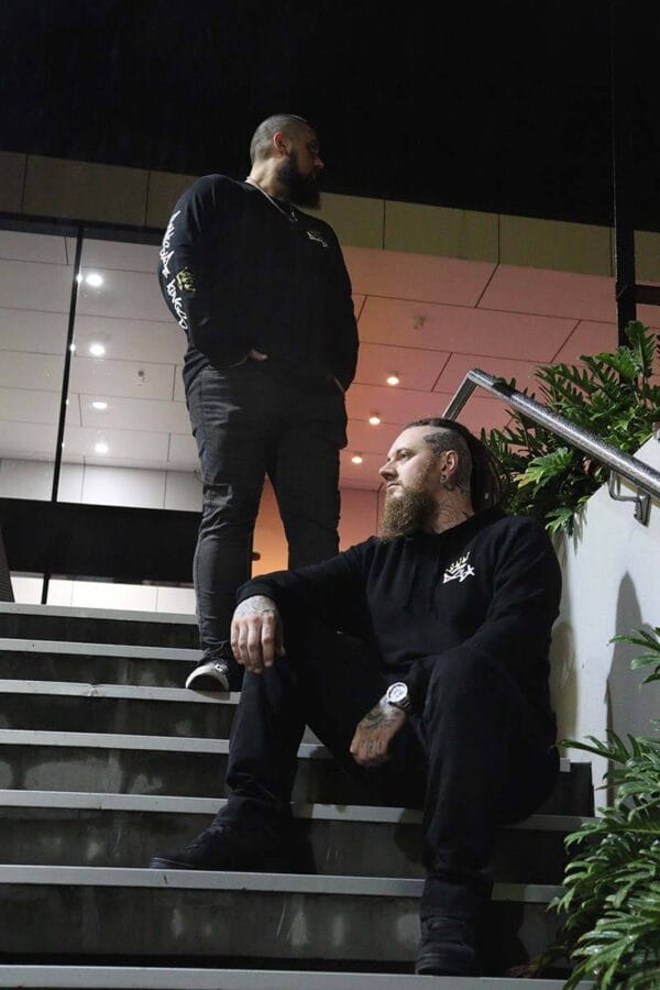 Male wearing a long sleeve DSK shirt standing on stairs while another male wearing a DSK hoodie sits on the lower stairs