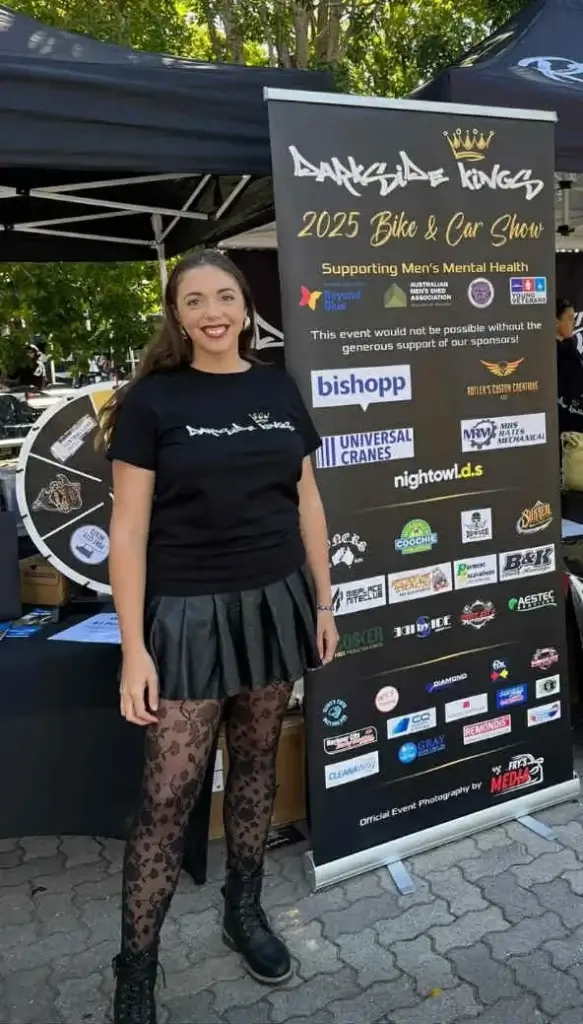 Lani Hilder standing in front of the 2025 Darkside Kings Clothing Bike & Car Show Sponsors Banner