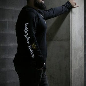 Male leaning against a concrete post wearing a black long sleeve shirt with a Darkside Kings logo printed down the sleeve