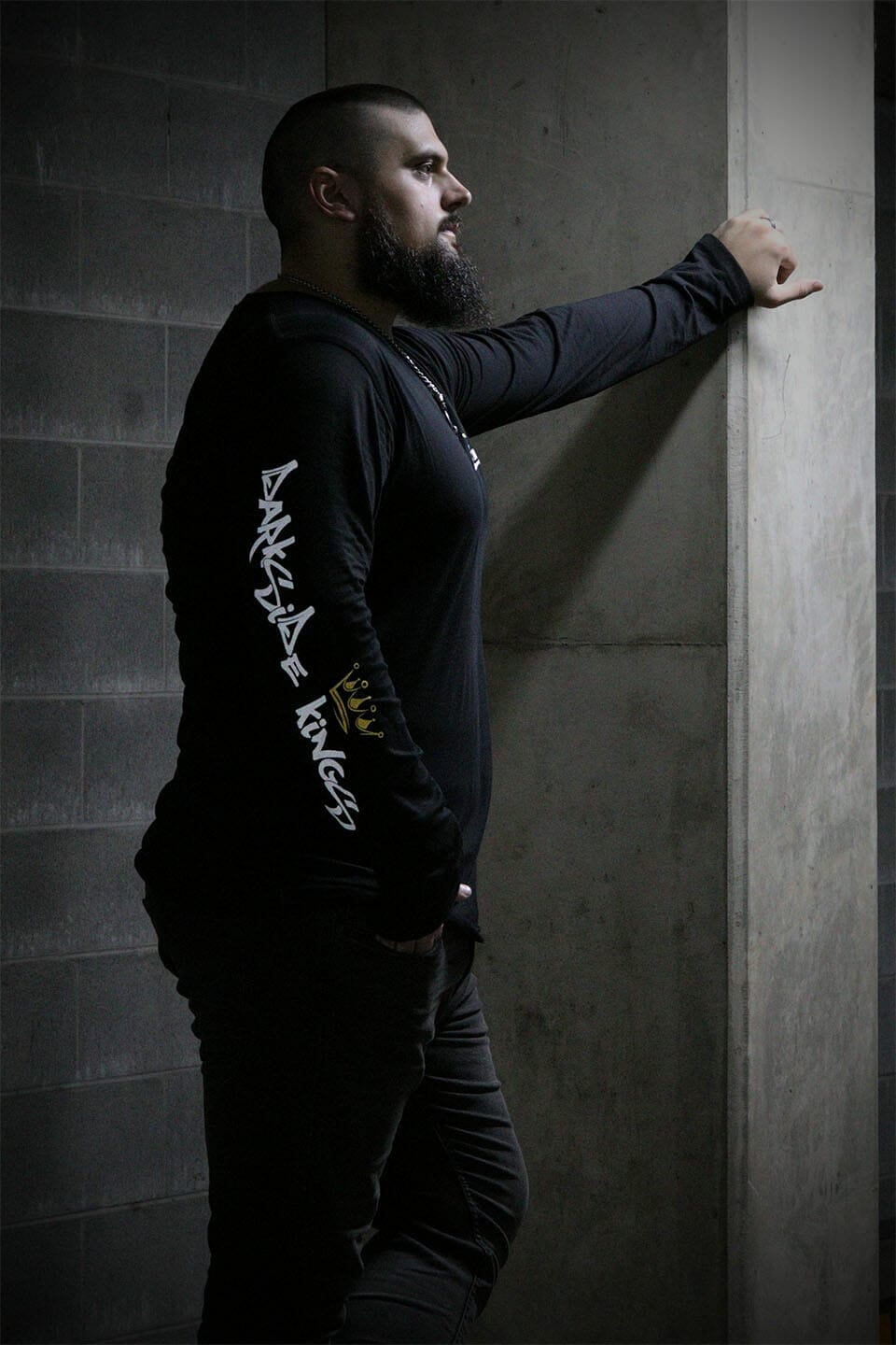 Male leaning against a concrete post wearing a black long sleeve shirt with a Darkside Kings logo printed down the sleeve