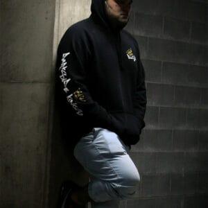 Male leaning against a concrete wall wearing a black hoodie with a Darkside Kings Logo printed down the sleeve and a DSK logo printed on the front