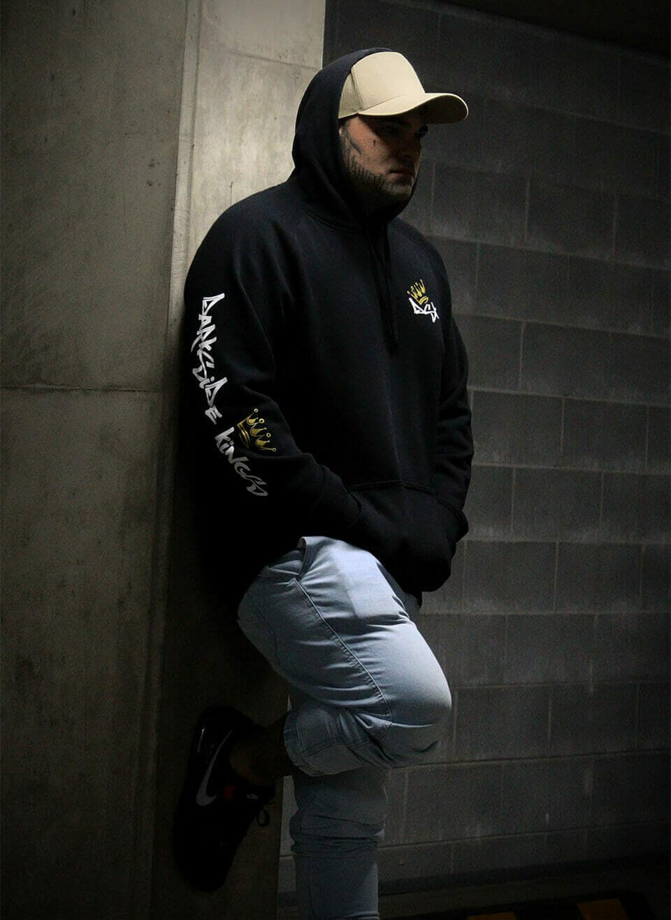 Male leaning against a concrete wall wearing a black hoodie with a Darkside Kings Logo printed down the sleeve and a DSK logo printed on the front