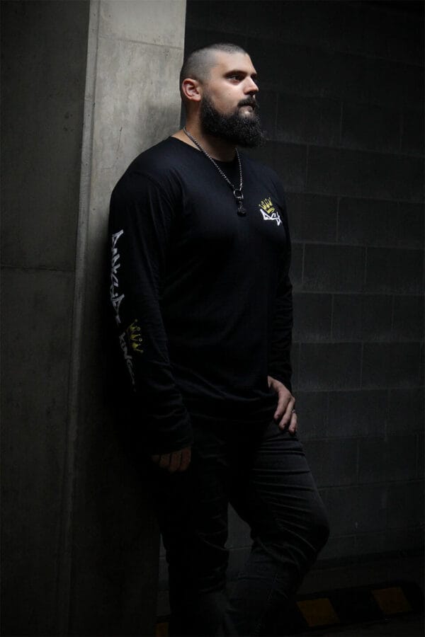 Male leaning against a concrete post wearing a black long sleeve shirt with a Darkside Kings logo printed down the sleeve and a DSK logo on the front