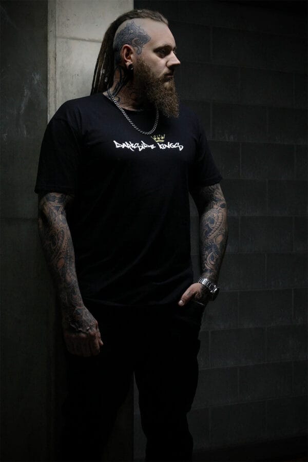 Male leaning against a concrete wall wearing a black Darkside Kings logo t-shirt